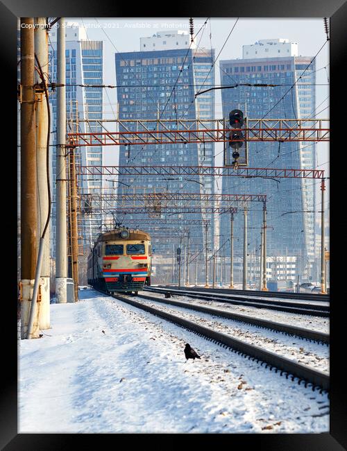 An electric train moves on rails against the backdrop of a cityscape of skyscrapers in a winter haze. Framed Print by Sergii Petruk