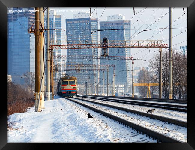 An old electric train moves on rails against the backdrop of a cityscape of skyscrapers on a sunny winter day. Framed Print by Sergii Petruk