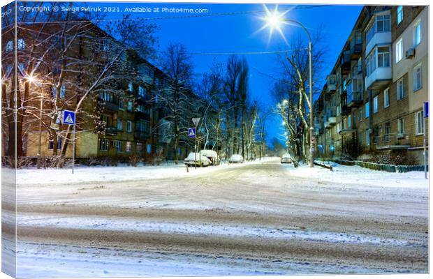 Snow-covered intersection on the road along a city street with trees in the snow and evening city illumination against the background of blue twilight. Canvas Print by Sergii Petruk