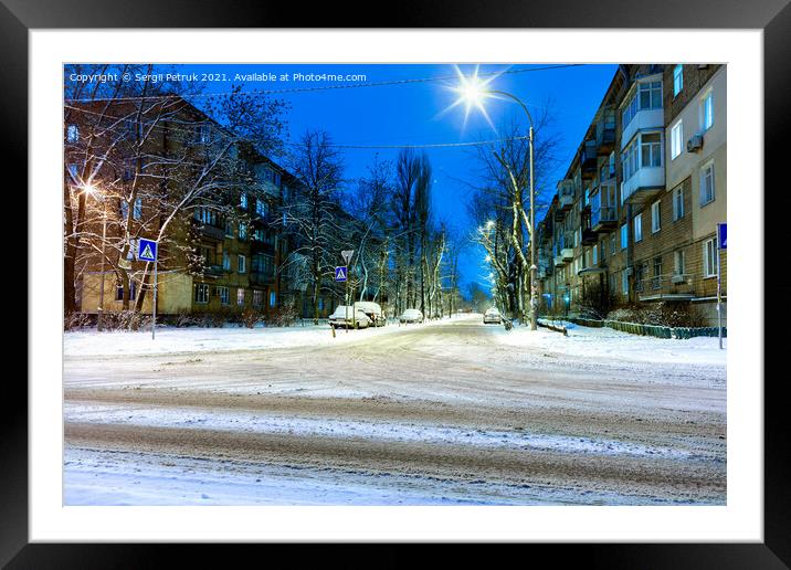 Snow-covered intersection on the road along a city street with trees in the snow and evening city illumination against the background of blue twilight. Framed Mounted Print by Sergii Petruk