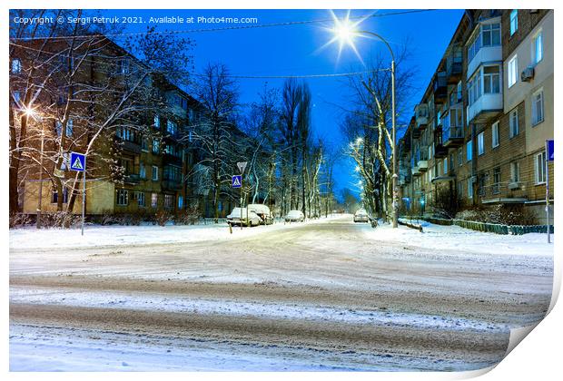 Snow-covered intersection on the road along a city street with trees in the snow and evening city illumination against the background of blue twilight. Print by Sergii Petruk