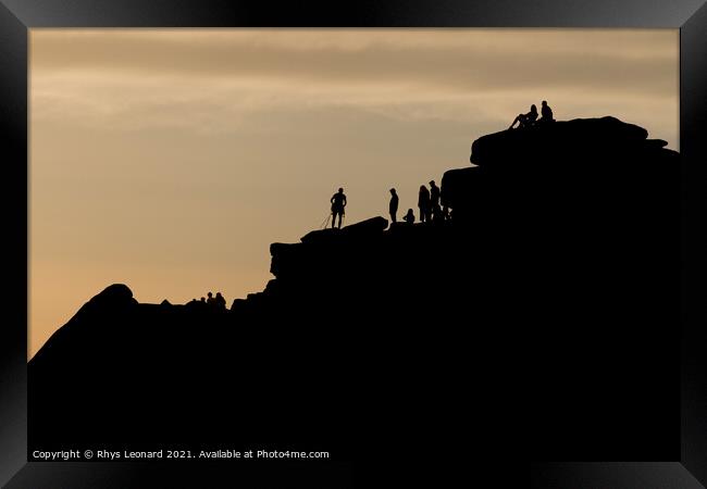 Silhouette of lots of people watching a climber at the summit of stanage edge. Climber wears a harness with tools like nuts, ropes and carabiners. Framed Print by Rhys Leonard