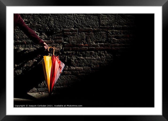 A beautiful colored umbrella rests folded against a brick wall i Framed Mounted Print by Joaquin Corbalan