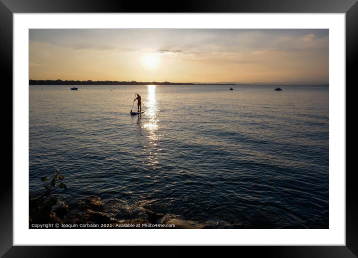A young man on a paddle surf board approaches the shore of the l Framed Mounted Print by Joaquin Corbalan