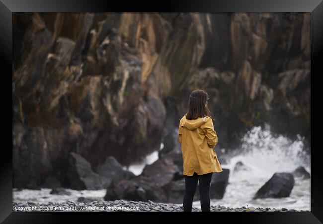 Woman on a yellow rain jacket looking to the sea with rocks on the background, in Madeira Framed Print by Luis Pina