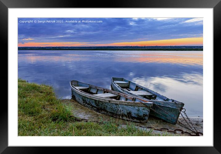 Two old dilapidated boats are moored with a metal chain to the bank of a calm river in the early morning against the backdrop of a cloudy sky and the rising sun. Framed Mounted Print by Sergii Petruk