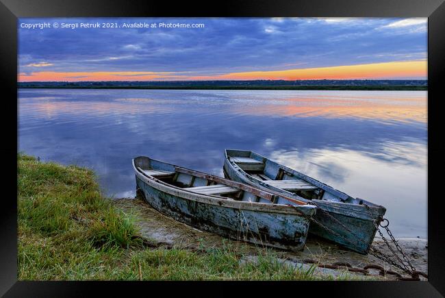 Two old dilapidated boats are moored with a metal chain to the bank of a calm river in the early morning against the backdrop of a cloudy sky and the rising sun. Framed Print by Sergii Petruk