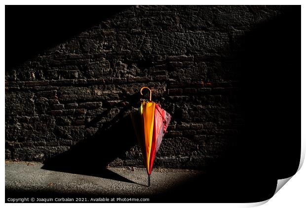 A beautiful colored umbrella rests folded against a brick wall i Print by Joaquin Corbalan