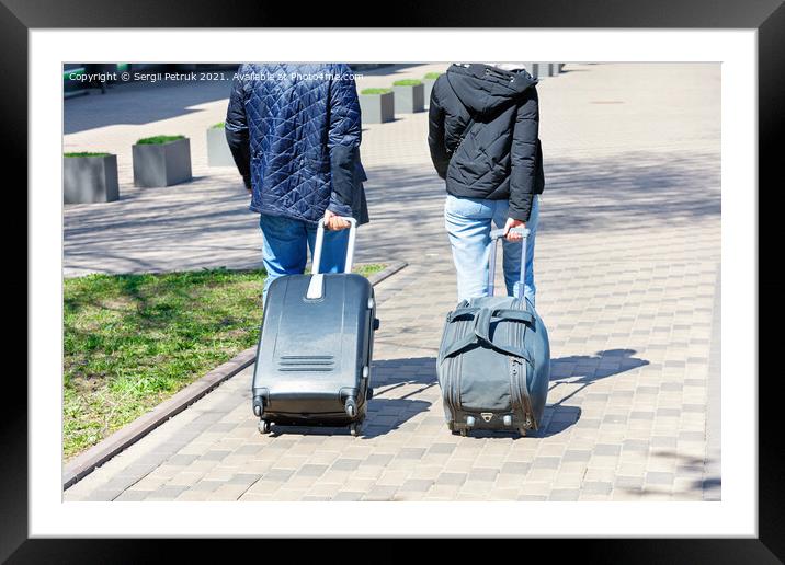 Two people, a man and a woman, walk along the cobbled sidewalk, carrying a travel bag and a suitcase on wheels behind them. Framed Mounted Print by Sergii Petruk