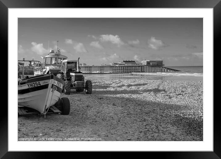 Black and white vintage photo of fishing boats and equipment on Cromer beach on the North Norfolk Coast. In the distance is the Victorian era pier Framed Mounted Print by Chris Yaxley