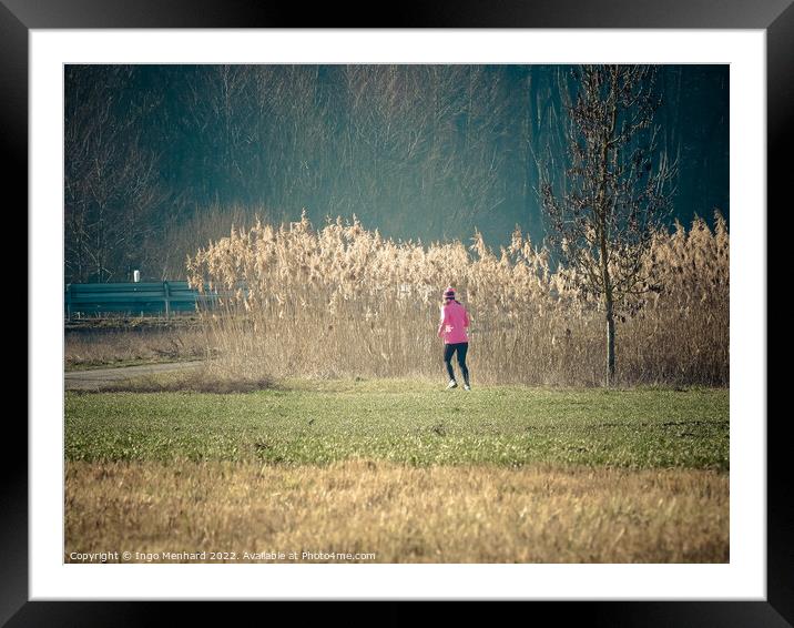 Woman in a pink jacket jogging in the park with dried reed grass background Framed Mounted Print by Ingo Menhard