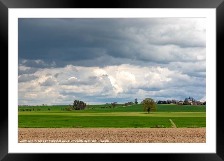 A small European village with a church on a hill, among spring fields, under a dramatic cloudy sky. Framed Mounted Print by Sergey Fedoskin