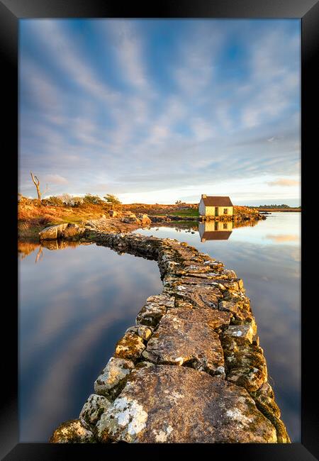 An old stone jetty across a lake leading to a small cottage Framed Print by Helen Hotson