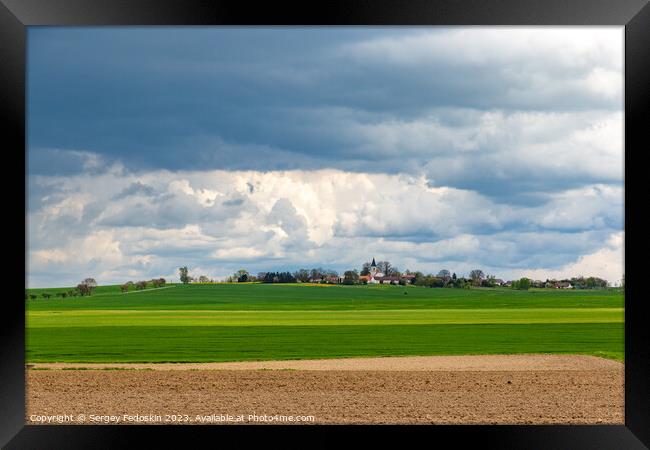 A small European village with a church on a hill, among spring fields, under a dramatic cloudy sky. Framed Print by Sergey Fedoskin