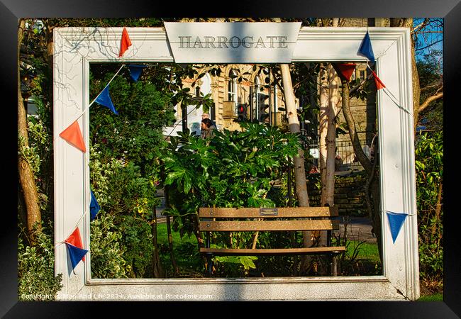 Quaint garden scene framed by a white wooden structure with 'Harrogate' sign, featuring a bench and lush greenery, adorned with colorful pennants. Framed Print by Man And Life