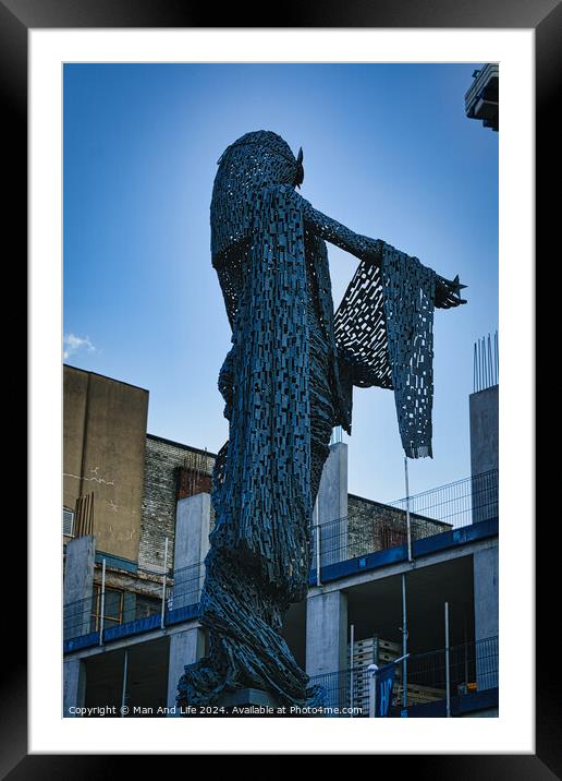 Artistic metal sculpture of a humanoid figure against a clear blue sky, with urban buildings in the background in Leeds, UK. Framed Mounted Print by Man And Life