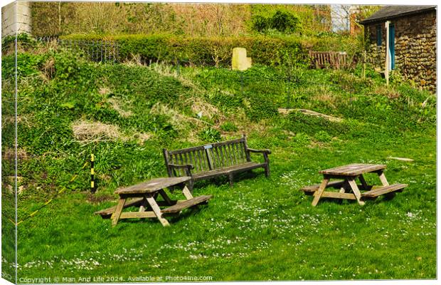 Rustic wooden benches and a table on a lush green grassy hillside, with a stone building and vegetation in the background, depicting a serene outdoor setting in York, North Yorkshire, England. Canvas Print by Man And Life
