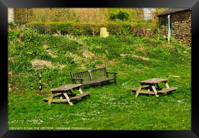 Rustic wooden benches and a table on a lush green grassy hillside, with a stone building and vegetation in the background, depicting a serene outdoor setting in York, North Yorkshire, England. Framed Print by Man And Life
