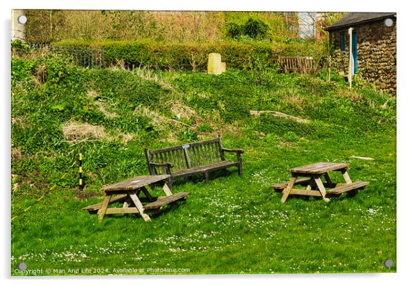 Rustic wooden benches and a table on a lush green grassy hillside, with a stone building and vegetation in the background, depicting a serene outdoor setting in York, North Yorkshire, England. Acrylic by Man And Life