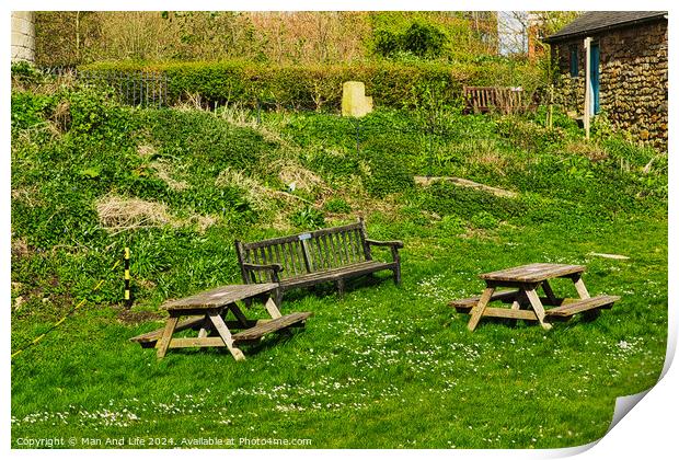 Rustic wooden benches and a table on a lush green grassy hillside, with a stone building and vegetation in the background, depicting a serene outdoor setting in York, North Yorkshire, England. Print by Man And Life