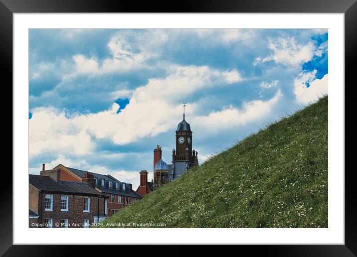 Quaint European town with historic buildings and a clock tower, set against a vibrant blue sky with fluffy clouds, and a lush green hill in the foreground in York, North Yorkshire, England. Framed Mounted Print by Man And Life