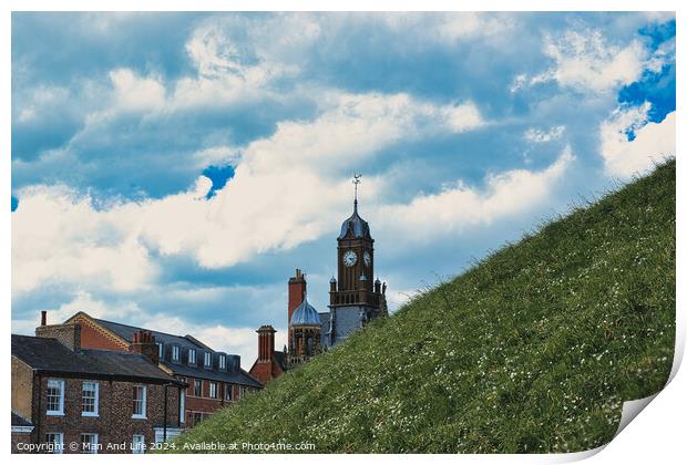 Quaint European town with historic buildings and a clock tower, set against a vibrant blue sky with fluffy clouds, and a lush green hill in the foreground in York, North Yorkshire, England. Print by Man And Life