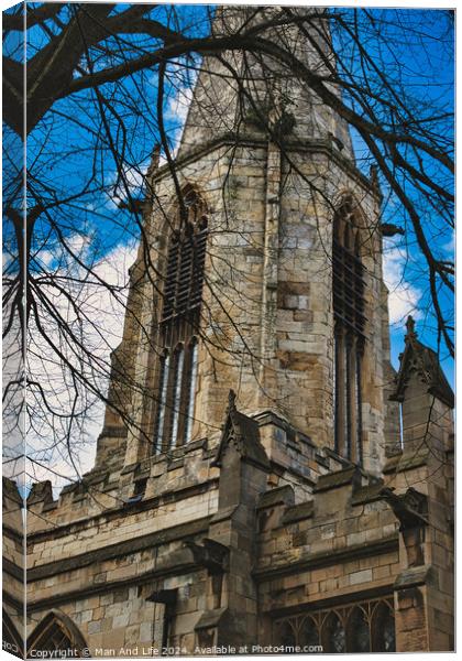 Gothic church tower with intricate stone details, framed by bare tree branches against a blue sky with fluffy clouds in York, North Yorkshire, England. Canvas Print by Man And Life