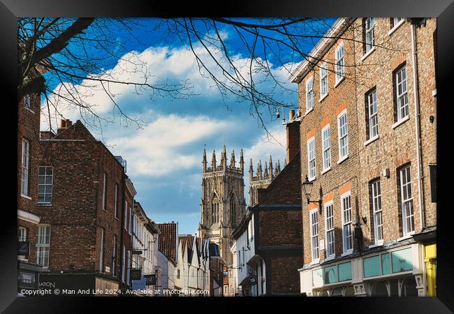 Historic European city street with traditional brick buildings and a prominent Gothic cathedral in the background under a blue sky with clouds in York, North Yorkshire, England. Framed Print by Man And Life