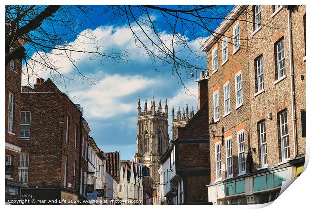 Historic European city street with traditional brick buildings and a prominent Gothic cathedral in the background under a blue sky with clouds in York, North Yorkshire, England. Print by Man And Life