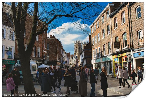Bustling city street scene with pedestrians, historic buildings, and a cathedral spire under a blue sky with scattered clouds in York, North Yorkshire, England. Print by Man And Life