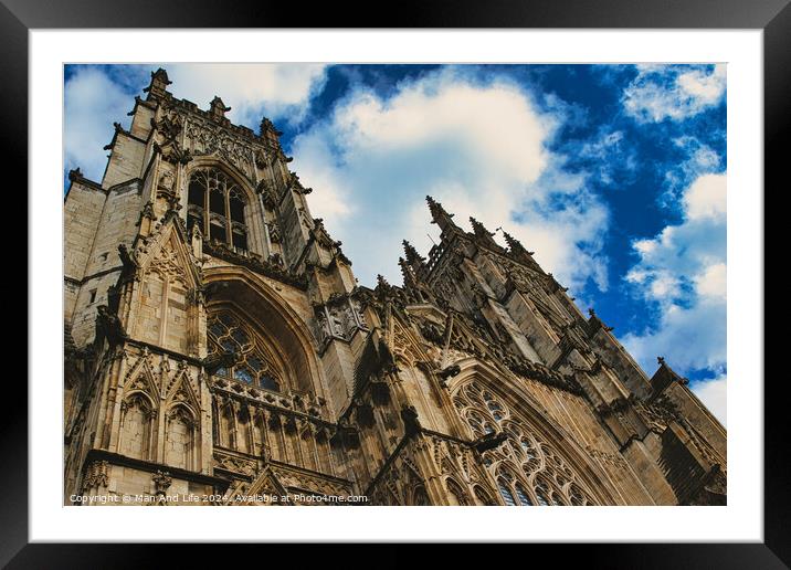 Dramatic angle of a Gothic cathedral's facade with intricate stone carvings against a vivid blue sky with fluffy clouds, showcasing architectural grandeur and historical elegance in York, North Yorkshire, England. Framed Mounted Print by Man And Life