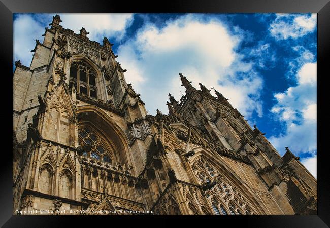Dramatic angle of a Gothic cathedral's facade with intricate stone carvings against a vivid blue sky with fluffy clouds, showcasing architectural grandeur and historical elegance in York, North Yorkshire, England. Framed Print by Man And Life