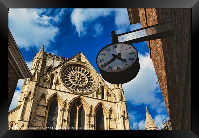 Vintage street clock hanging with a gothic cathedral facade in the background, showcasing intricate architecture and a clear blue sky in York, North Yorkshire, England. Framed Print by Man And Life