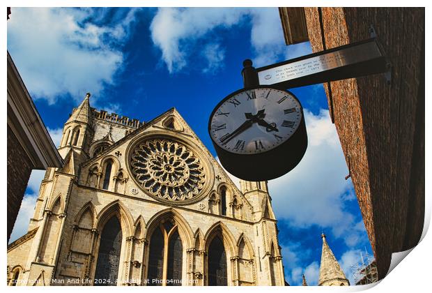 Vintage street clock hanging with a gothic cathedral facade in the background, showcasing intricate architecture and a clear blue sky in York, North Yorkshire, England. Print by Man And Life