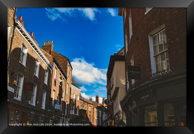 Charming European street scene with historic brick buildings under a clear blue sky with fluffy clouds, showcasing architectural details and local businesses in York, North Yorkshire, England. Framed Print by Man And Life