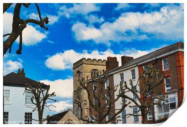 Quaint urban scene with historic stone tower, traditional buildings, and bare tree branches against a vibrant blue sky with fluffy white clouds in York, North Yorkshire, England. Print by Man And Life