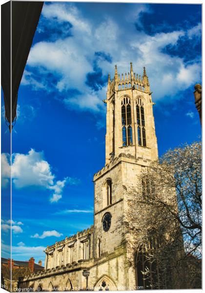 Gothic church tower against a vibrant blue sky with fluffy clouds, showcasing intricate architectural details and a blooming tree at the corner in York, North Yorkshire, England. Canvas Print by Man And Life