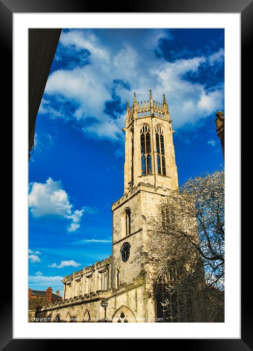 Gothic church tower against a vibrant blue sky with fluffy clouds, showcasing intricate architectural details and a blooming tree at the corner in York, North Yorkshire, England. Framed Mounted Print by Man And Life