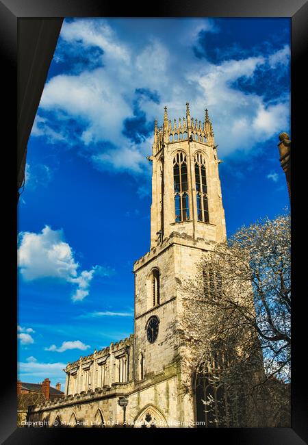 Gothic church tower against a vibrant blue sky with fluffy clouds, showcasing intricate architectural details and a blooming tree at the corner in York, North Yorkshire, England. Framed Print by Man And Life