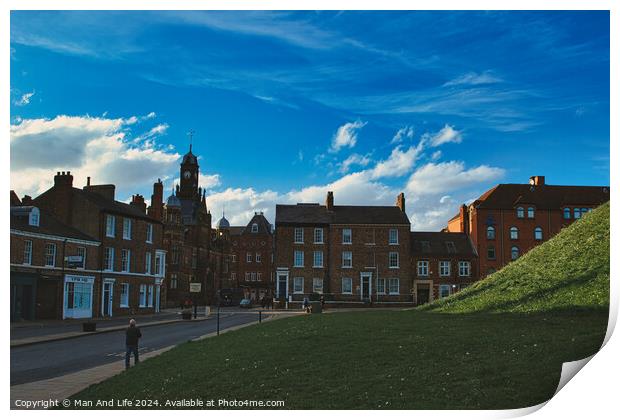Quaint European town with historic buildings under a dramatic blue sky with fluffy clouds, featuring a lush green hill and pedestrians on a sidewalk in York, North Yorkshire, England. Print by Man And Life