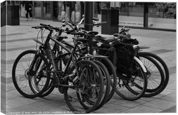 Black and white image of multiple bicycles locked to a bike rack in an urban setting, with a blurred background of a city street Canvas Print by Man And Life