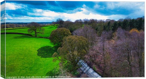 Aerial view of a vibrant countryside landscape with lush green fields, a meandering road, and a patch of bare trees under a dynamic sky with scattered clouds. Canvas Print by Man And Life