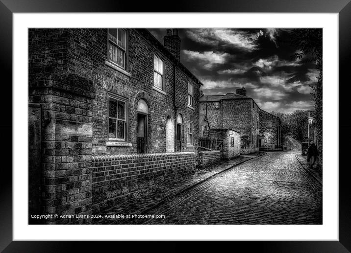Victorian cobble stone street and shops England lit by lamps  Framed Mounted Print by Adrian Evans