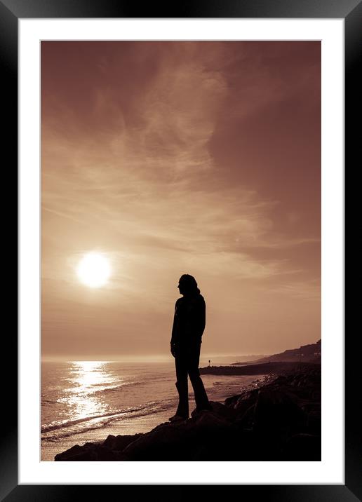 Silhouetted girl stands by sea at sunset Framed Mounted Print by George Cairns