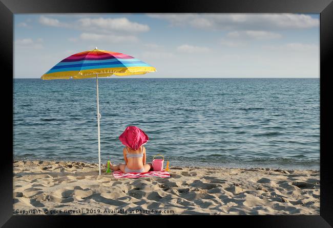 little girl with hat sitting under sunshade on bea Framed Print by goce risteski