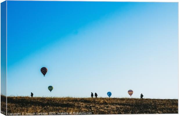 Silhouettes of tourists walking through a meadow while watching hot air balloons flying on the horizon, blue sky background, copy space, added film grain. Canvas Print by Joaquin Corbalan