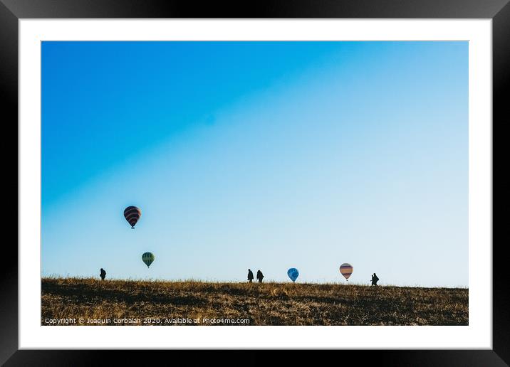 Silhouettes of tourists walking through a meadow while watching hot air balloons flying on the horizon, blue sky background, copy space, added film grain. Framed Mounted Print by Joaquin Corbalan