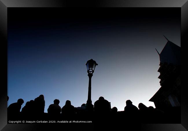  A crowd of tourists rests at dusk on a bridge, silhouetted images backlight against a streetlight. Framed Print by Joaquin Corbalan