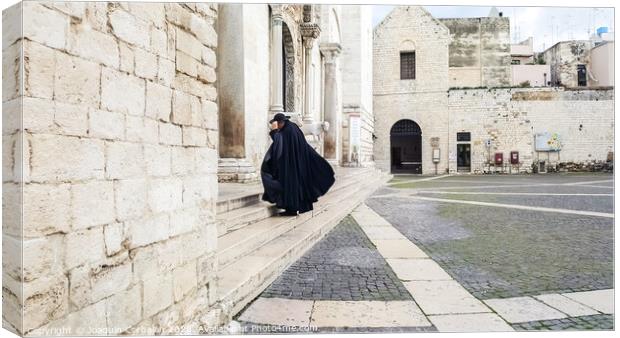 Bari, Italy - March 12, 2019: A Catholic priest walks towards the cathedral dressed in a black cape and hat a day of wind and rain. Canvas Print by Joaquin Corbalan