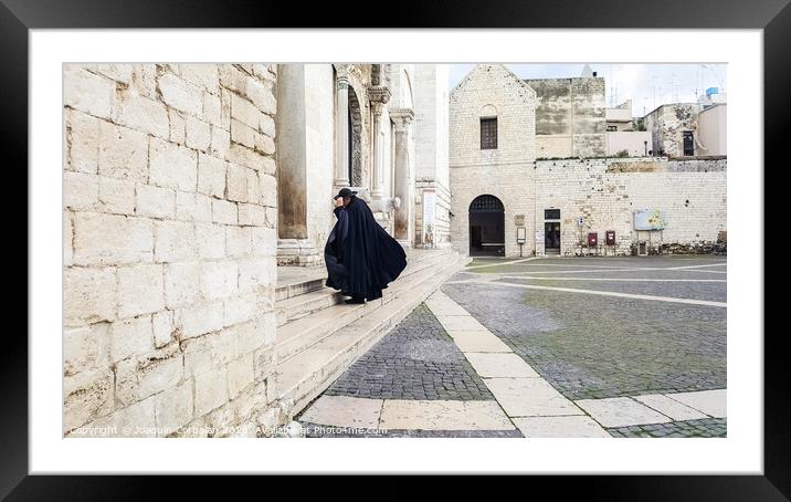 Bari, Italy - March 12, 2019: A Catholic priest walks towards the cathedral dressed in a black cape and hat a day of wind and rain. Framed Mounted Print by Joaquin Corbalan